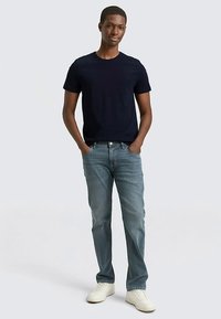 Navy blue short-sleeve t-shirt paired with light blue jeans and white sneakers. Model stands with hands in pockets against a plain background.