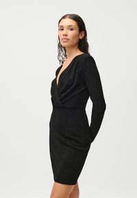 Μαύρο φόρεμα με μακριά μανίκια και βαθύ V-ντεκολτέ. Φτιαγμένο από υφασμάτινη υφή που γυαλίζει. Στενή σιλουέτα με λεπτομέρεια στροφής στη μέση.