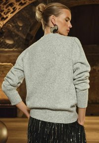 Jasnoszara dziergana sweter z teksturowanym wykończeniem, zebranymi ramionami i prążkowanymi mankietami. Połączony z czarną spódnicą z frędzlami.
