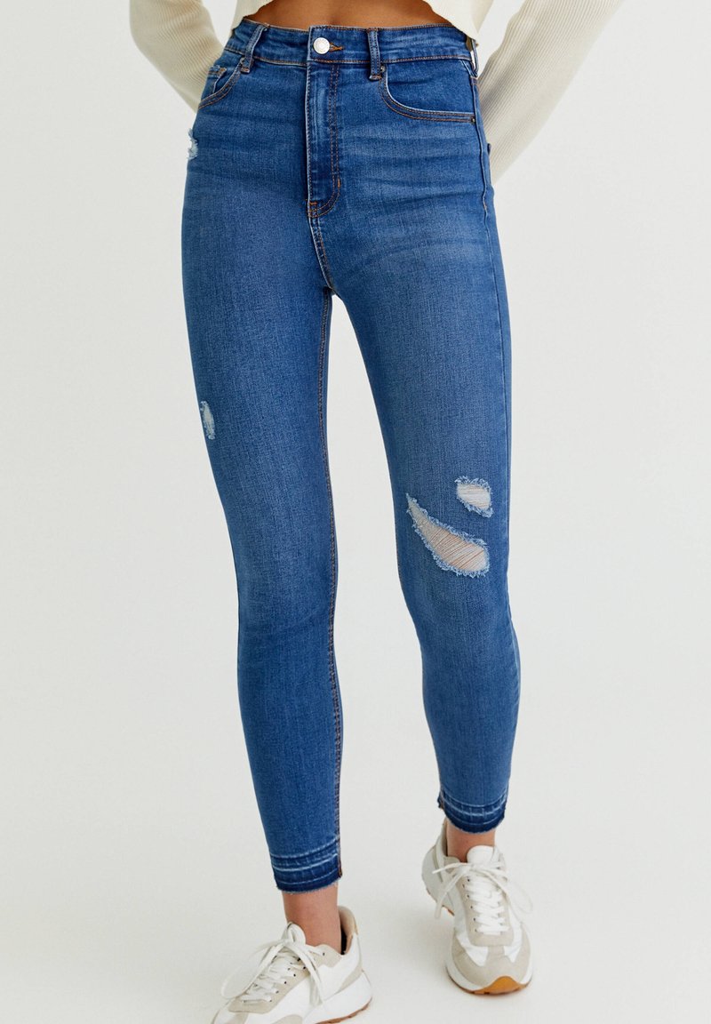 Tunna blå denimjeans med hög midja, med slitna hål på knäna och en ren nederkant. I kombination med vita och beige sneakers.