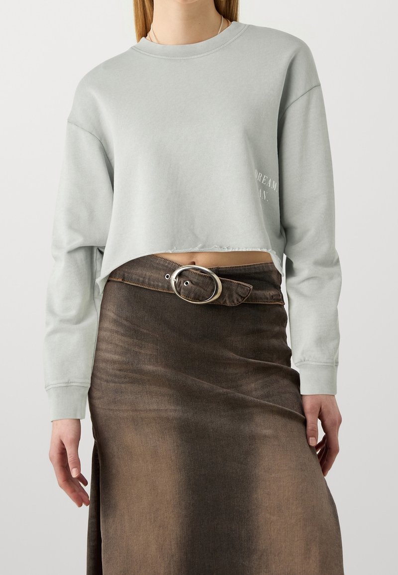Femme portant un sweat-shirt court gris clair à manches longues et une jupe en denim marron délavé avec une ceinture asymétrique à boucle ovale argentée.
