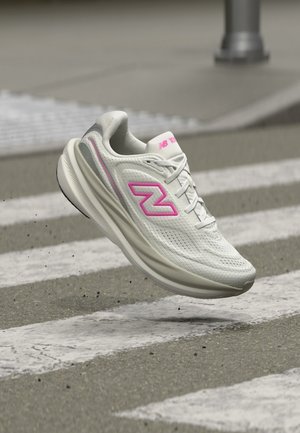 Chaussure de running New Balance blanche avec logo rose, en plein vol au-dessus d'un passage piéton sur une route asphaltée.