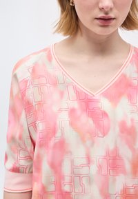 Γυναίκα που φοράει ροζ και λευκή μπλούζα tie-dye με γεωμετρικά περιγράμματα και λαιμόκοψη V, με κοντά ξανθά μαλλιά και χρυσούς κρίκους στα αυτιά.