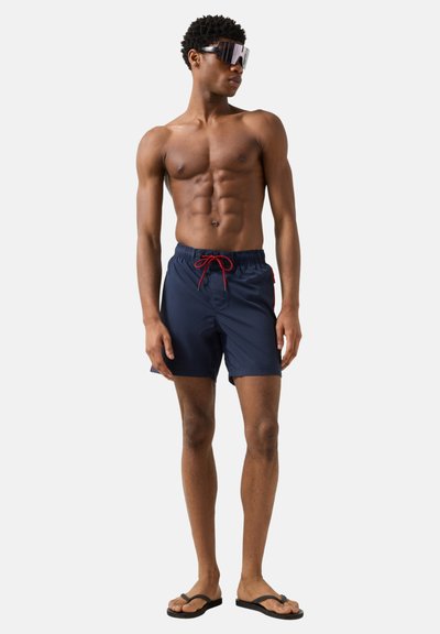 Homme torse nu portant un short de bain bleu marine avec un cordon rouge, des tongs noires et de grandes lunettes de soleil enveloppantes, debout devant un fond blanc.
