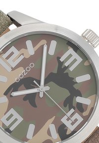 Montre analogique avec un boîtier rond en argent et un cadran à motifs camouflage. Présente de grands index d'heure en relief et un bracelet en tissu vert texturé.