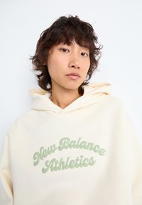 Jeune personne avec des cheveux courts et bouclés portant un sweat à capuche crème avec le texte "New Balance Athletics" en vert clair à l'avant.