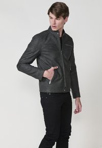 Giacca in pelle grigia scura con colletto rialzato, chiusura a zip sul davanti, tasche sul petto e ai lati, dettagli a coste sulle spalle e una vestibilità aderente.