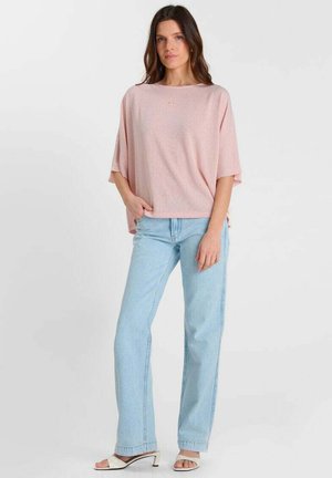 Femme debout portant un pull rose ample, un jean droit bleu clair et des sandales blanches à talons et bouts ouverts sur un fond uni.