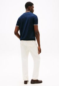 Camicia navy a maniche corte con polsini a strisce bianche, abbinata a pantaloni bianchi e scarpe in pelle marrone, vista da dietro. Texture del tessuto liscia.