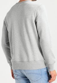 Hombre vestido con sudadera gris claro sencilla y jeans azul claro, visto desde atrás, de pie frente a un fondo blanco.