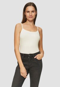Vrouw met lang bruin haar, gekleed in een witte top met spaghetti-bandjes en hooggewaist zwarte jeans, poseert met één hand in de zak.