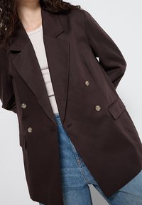 Femme portant un blazer croisé marron foncé sur un haut blanc et un jean bleu, debout devant un fond uni clair.