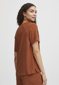 Top a maniche corte color ruggine realizzato in tessuto leggero con una vestibilità rilassata e un orlo spaccato. Presenta un collo rotondo e una trama sottile.