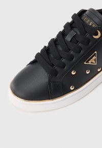 Sneaker in pelle nera con lacci neri, dettagli con borchie dorate, rifinitura dorata sulla suola bianca e logo Guess dorato sul lato e sulla linguetta.