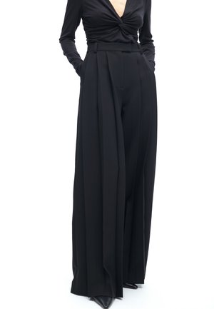 Black wide-leg trousers made from a smooth material, featuring pleats, a high waistband, and side pockets. Paired with a deep V-neck top.