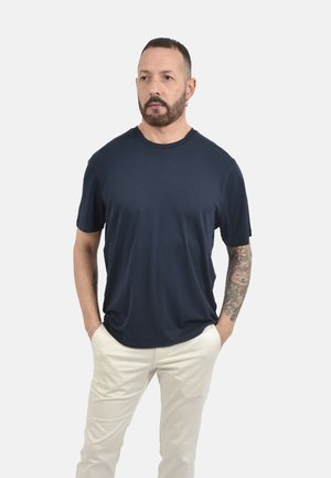 Uomo con il braccio destro tatuato e la barba, che indossa una maglietta blu navy e pantaloni chiari, in piedi con le mani nelle tasche.