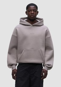 Felpa con cappuccio di un grigio chiaro, dotata di tasca frontale, polsini larghi e un cappuccio ampio. Presenta un logo minimale sul petto. Tessuto morbido e liscio.