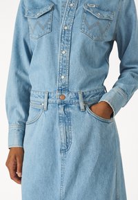  világoskék farmerruha, hosszú ujjakkal, két mellzsebbel és övvel a derékon. Jellegzetes varrási részletek adnak szövetérzetet.