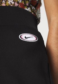 Svart ribbad kjol med en cirkulär broderad Nike-logotyp i rosa och vitt på midjan. Tyget har en strukturerad yta.