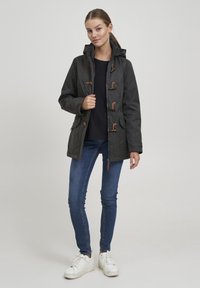 Mørk grå hettegenser med tretoggle-lukking, med sidelommer og glidelås, brukt over en svart topp og blå jeans.