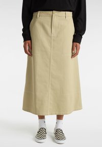 Ljusbeige denimkjol med vertikal sömnad och synliga fickor, åtföljd av svarta och vita rutiga slip-on skor och vita strumpor.