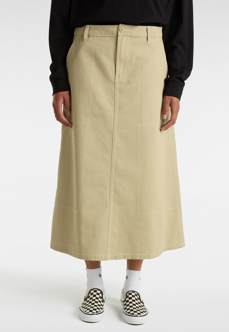 Ljusbeige denimkjol med vertikal sömnad och synliga fickor, åtföljd av svarta och vita rutiga slip-on skor och vita strumpor.