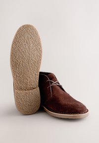 Braune Wildlederschuhe mit einer runden Spitze, hellfarbiger Sohle und strukturiertem Unterboden. Sie verfügen über graue Schnürsenkel und weiße Nähte entlang des Randes.