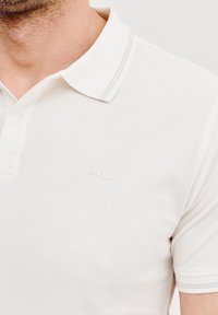Biała koszulka polo z teksturowanej tkaniny, klasyczny kołnierzyk z szarymi akcentami i małym logo na klatce piersiowej.