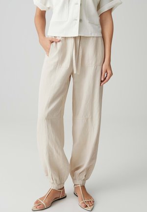 Femme portant un pantalon beige à cordon de serrage avec des poignets élastiques, une veste courte crème et des sandales plates beiges à lanières.