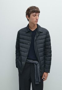 Schwarze steppierte Pufferjacke mit Knopfverschluss und Kragen. Trägt man über einem navyblauen Pullover, kombiniert mit dunklen Hosen und einem grauen Schal, der in der Taille gebunden ist.