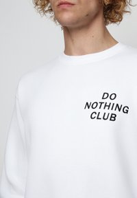 Bílá mikina vyrobená z bavlněné směsi s kulatým výstřihem. Na hrudi má vyšitý text "DO NOTHING CLUB" v tučném černém písmu.