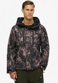 Man in een zwart en bruin geblokt hoodie-jack met meerdere ritssluitingen, staande tegen een effen witte achtergrond.