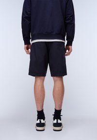 Persoon staande, gekleed in zwarte korte broek, marineblauwe trui, zwarte sokken en witte en zwarte sneakers op een effen lichte achtergrond.