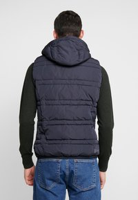 Marine-blaues Puffer-Weste mit Kapuze, ausgestattet mit vertikalen Nählinien und einer leicht glänzenden Textur. Getragen über einem dunkelgrünen Pullover.