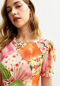 Blommönstrad lappklänning i nyanser av rosa, orange och grön, med puffärmar, insydd midja och rund halsringning. Guldskiktad halsband.