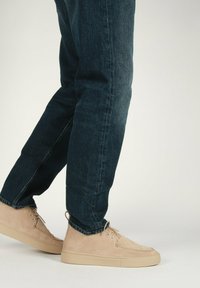 Sneaker alla caviglia in suede beige con suola piatta in gomma e lacci abbinati. Indossati con jeans in denim blu scuro, che mettono in evidenza un look casual.