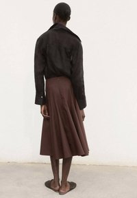 Schwarzes Leinenhemd mit langen Ärmeln und einem gebogenen Kragen, kombiniert mit einem braunen, plissierten Midirock. Flache, geflochtene Schuhe runden das Outfit ab.