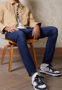 Beige bomullsjacka med dragkedja, blå jeans i denim och vita sneakers med marinblå detaljer. Trästolen har en enkel design.