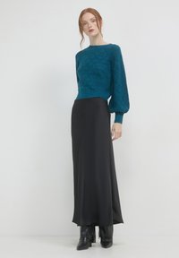 Frau mit roten Haaren, die einen strukturierter, türkisfarbenen Pullover mit Puffärmeln, einen langen schwarzen Rock und schwarze Stiefel trägt, vor einem schlichten weißen Hintergrund.