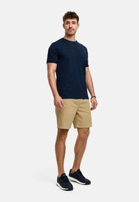 Mørkeblå kortærmet T-shirt med lomme; parret med beige shorts. Modellen har mørke sneakers på og står foran en ensfarvet baggrund.