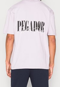 Osoba oblečená v svetle fialovém tričku s velkým černým nápisem "PEGADOR" a menším bílým kurzivním nápisem "Modern Reality" na zadní straně, doplněná tmavými kalhotami.