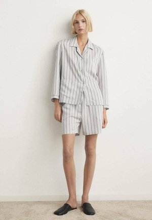 Mujer con cabello rubio corto que lleva una camisa de manga larga blanca a rayas y pantalones cortos a juego, de pie sobre una alfombra beige con zapatos negros sin cordones.