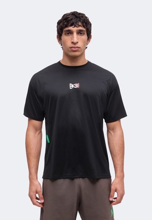 Homme aux cheveux courts et bouclés portant un t-shirt noir avec un petit logo blanc et rouge sur la poitrine et un pantalon marron, debout devant un fond uni.