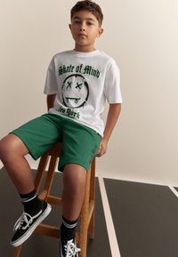 Wit T-shirt met groene grafische print, gecombineerd met groene shorts. Zwarte sneakers met witte veters en zwart-wit gestreepte sokken. Houten krukje.