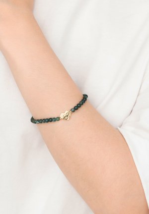 Arm mit dunkelgrünem Perlenarmband und goldenem, geometrischem Herzanhänger vor weißem Stoffhintergrund.