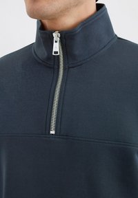 Marinblå zip-up tröja i mjukt tyg, med hög krage, silverfärgad dragkedja och horisontella sömmar över bröstet.
