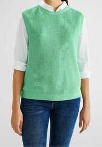Light green knitted sleeveless top with ribbed texture and round neckline, worn over a white collared shirt, paired with blue jeans.