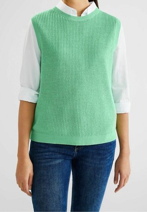  lichtgroene gebreide mouwloze top met geribbelde structuur en ronde hals, gedragen over een witte blouse met kraag, gecombineerd met blauwe jeans.