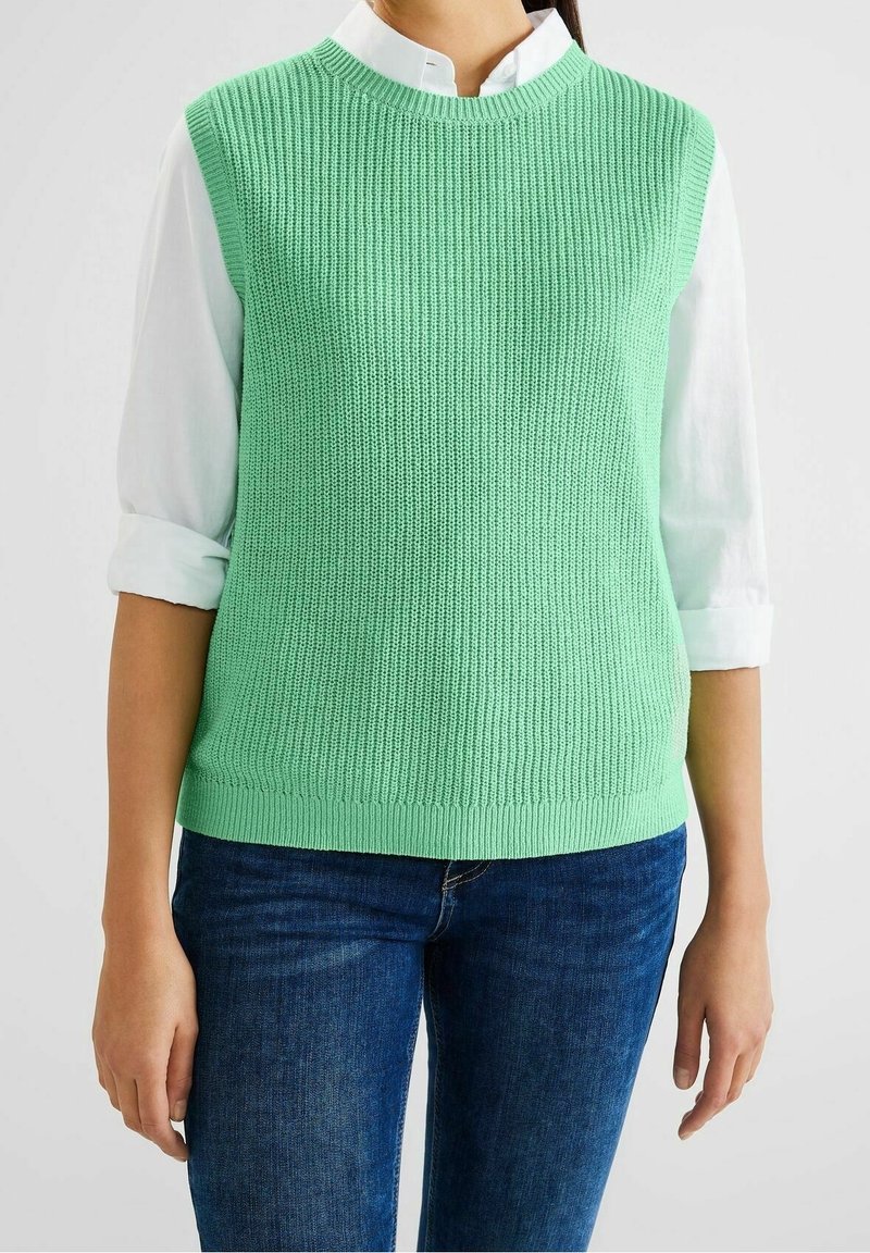 Light green knitted sleeveless top with ribbed texture and round neckline, worn over a white collared shirt, paired with blue jeans.