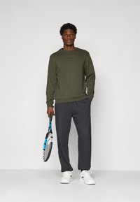 Bărbat îmbrăcat într-un hanorac verde măsliniu și pantaloni negri, ținând o rachetă de tenis neagră și albastră, stând pe un fundal deschis simplu.